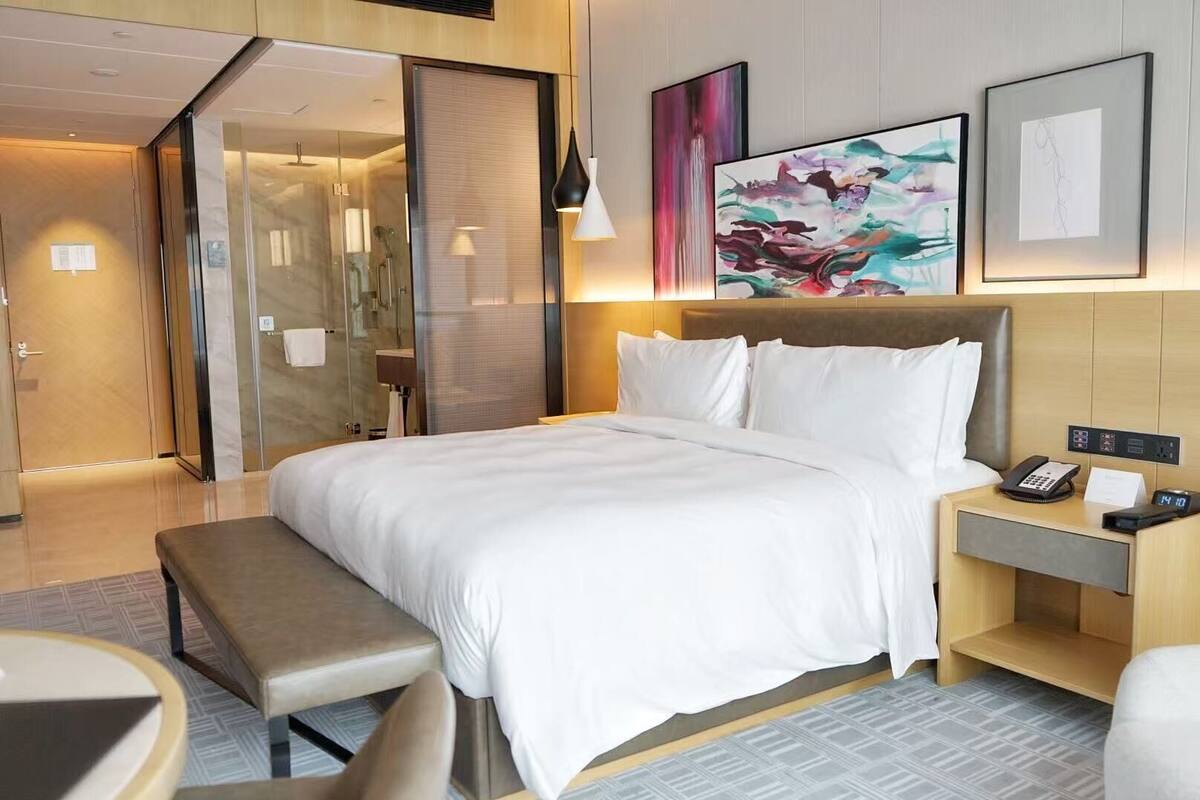 A hotel room with a bed, bedside table, lamp, artwork, and a bathroom visible through a glass door.