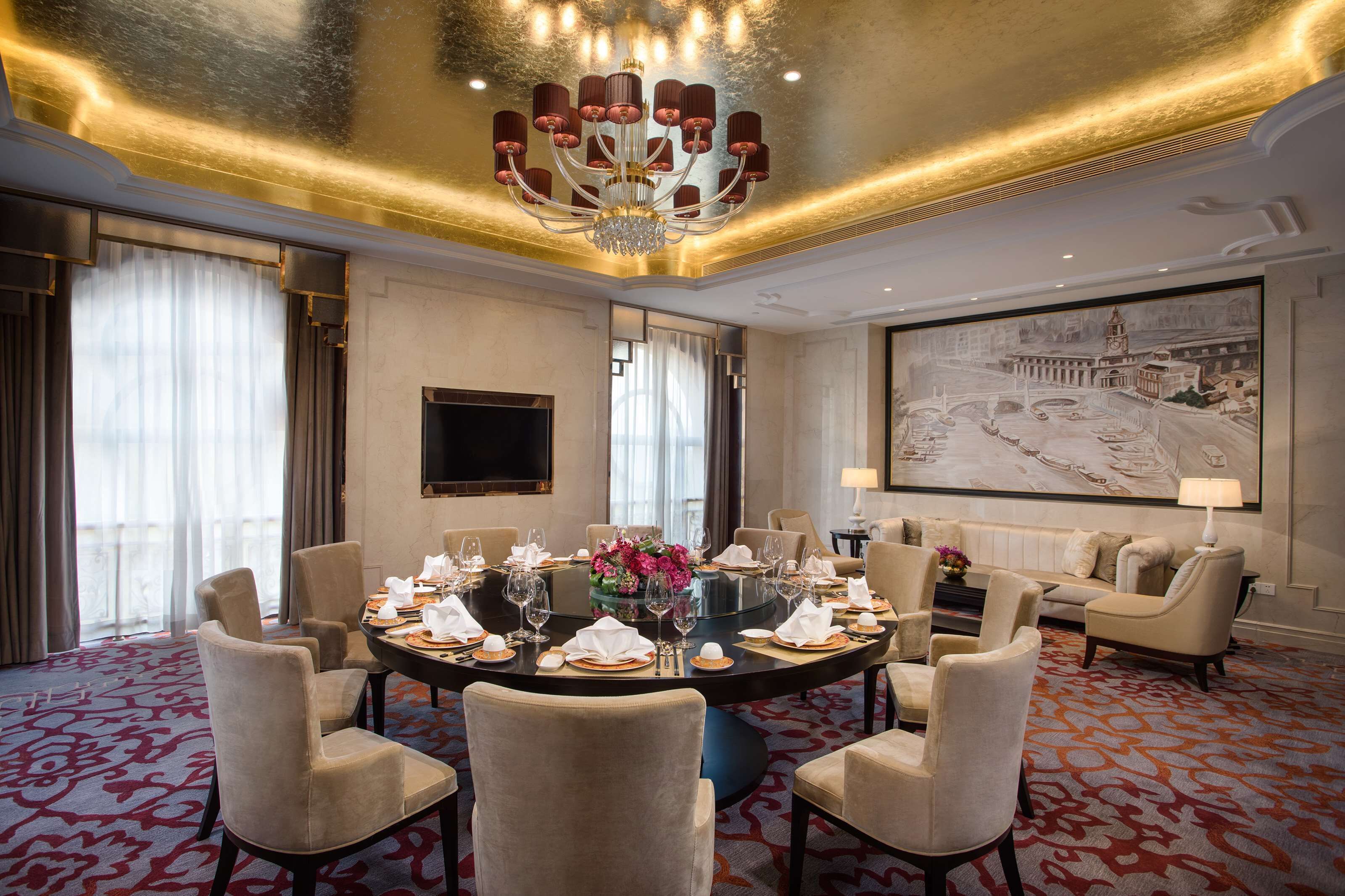 Un comedor de hotel con una mesa redonda dispuesta para seis personas, rodeada de seis sillas, un candelabro, un cuadro grande en la pared y un televisor montado.