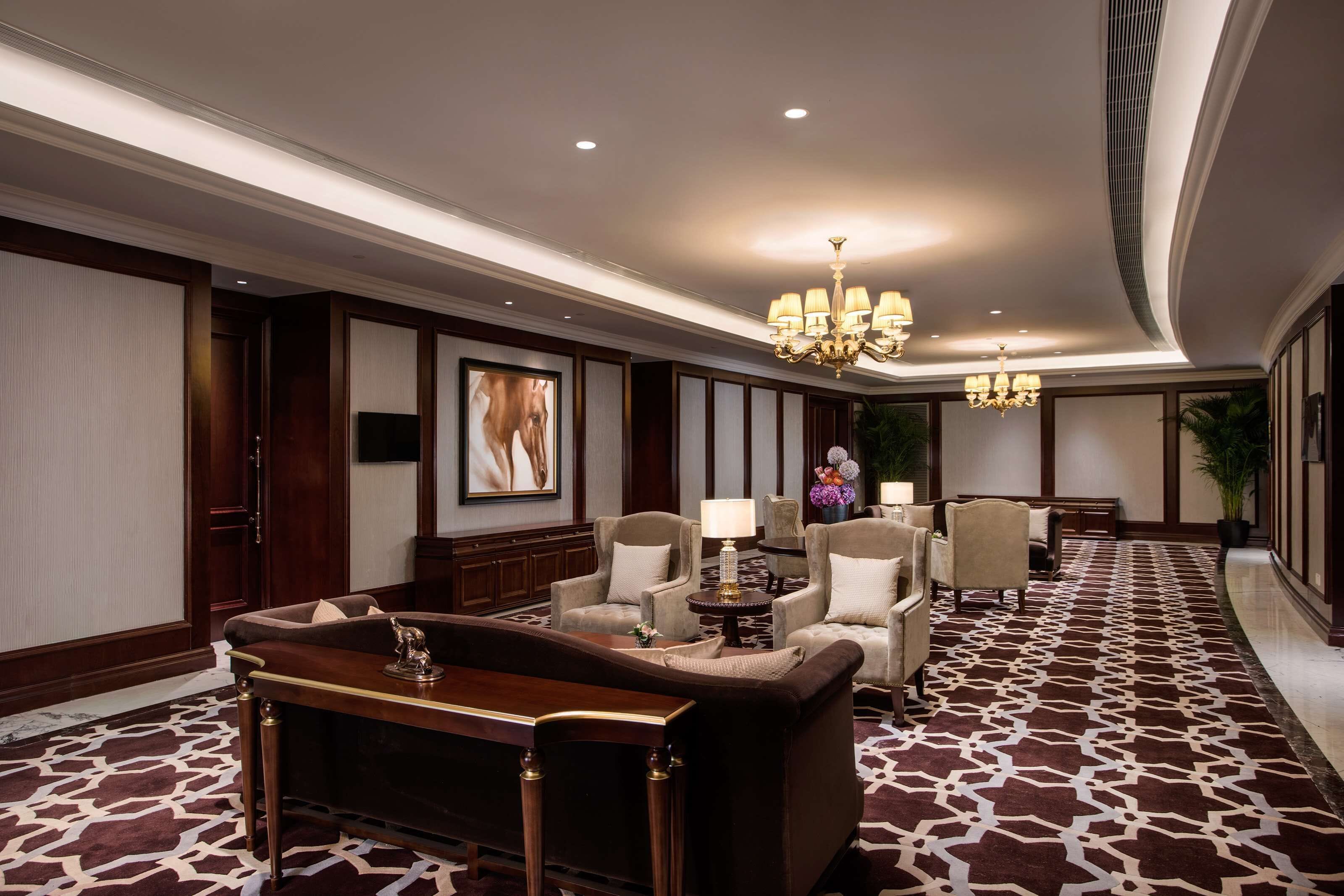A hotel lobby with a patterned carpet, a reception desk, several armchairs, a chandelier, and a large framed picture on the wall.