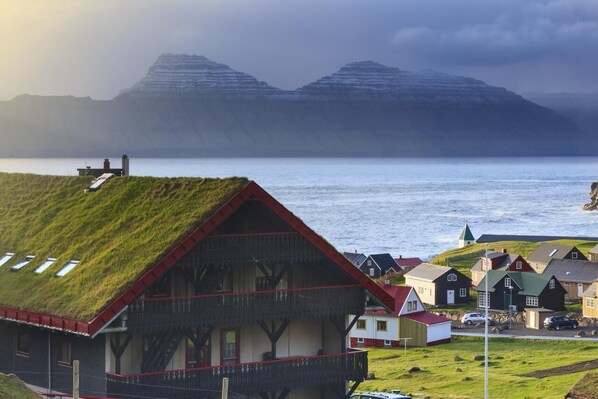 A traditional house with a grass roof, situated in a coastal village with houses in the background and mountains in the distance. - Gjaargardur Guesthouse Gjogv (Gjogv)