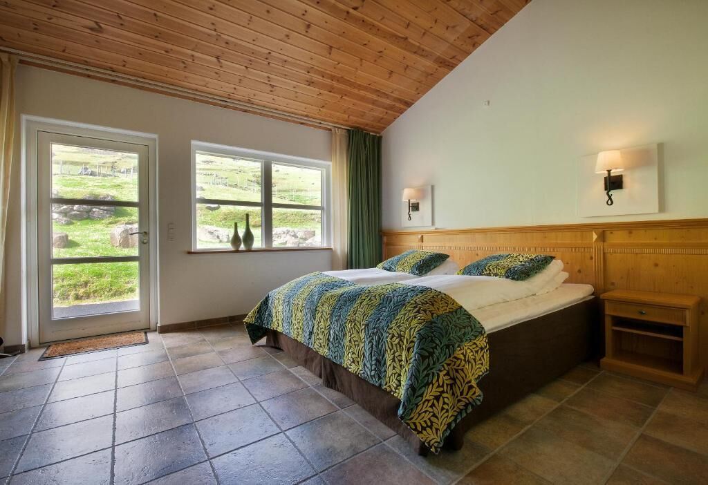 A bedroom with a large bed, wooden ceiling, two windows with curtains, and a bedside table.