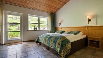 A bedroom with a large bed, wooden ceiling, two windows with curtains, and a bedside table.