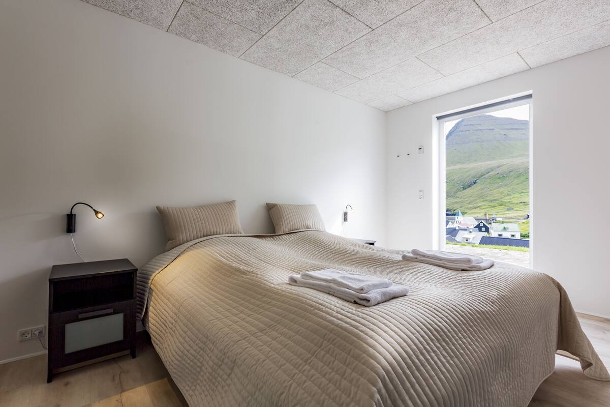 A bedroom with a large bed, a nightstand, a wall-mounted lamp, and a window with a view of a green landscape.
