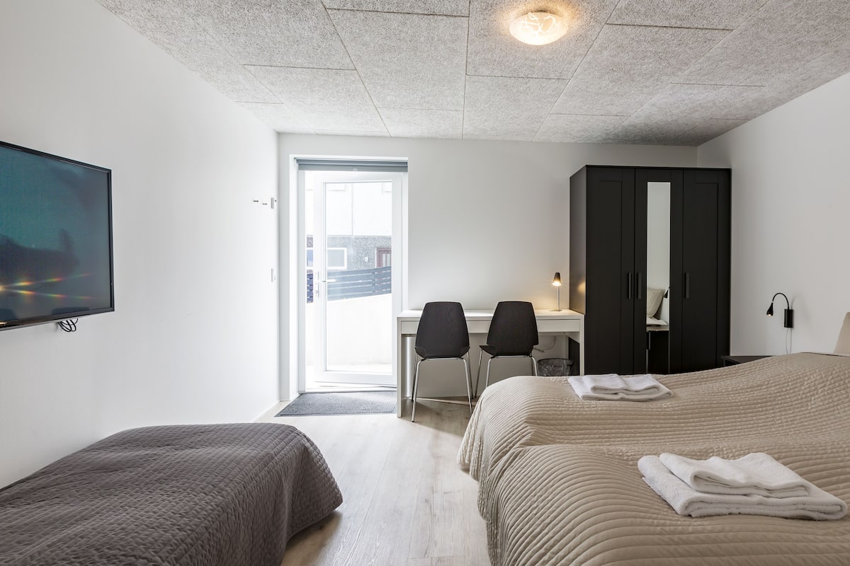 A modern hotel room with a large bed, a smaller bed, a desk with a chair, a wardrobe, and a television.