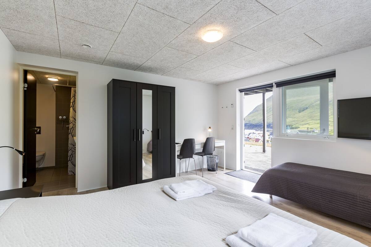 A modern bedroom with a large bed, a desk, and a chair. There is a bathroom visible through an open door.