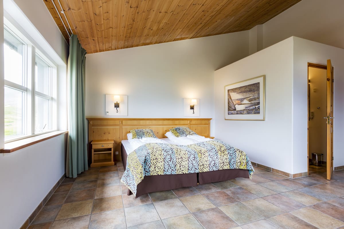 A bedroom with a wooden ceiling, a large bed with patterned bedding, bedside lamps, a nightstand, a window with curtains, and a painting on the wall.