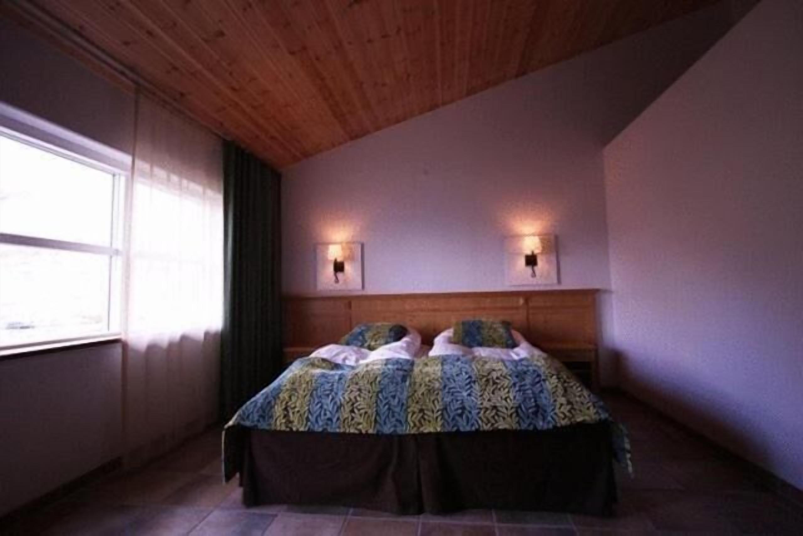 Une chambre avec un plafond en bois, un lit recouvert d’une couvre-lit à motifs, deux lampes fixées au mur et une fenêtre avec des rideaux.