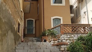 Eine Treppe aus Stein führt zu einem Gebäude mit heller Fassade, einem kleinen Balkon und einem Fenster mit verzierten Rollläden.