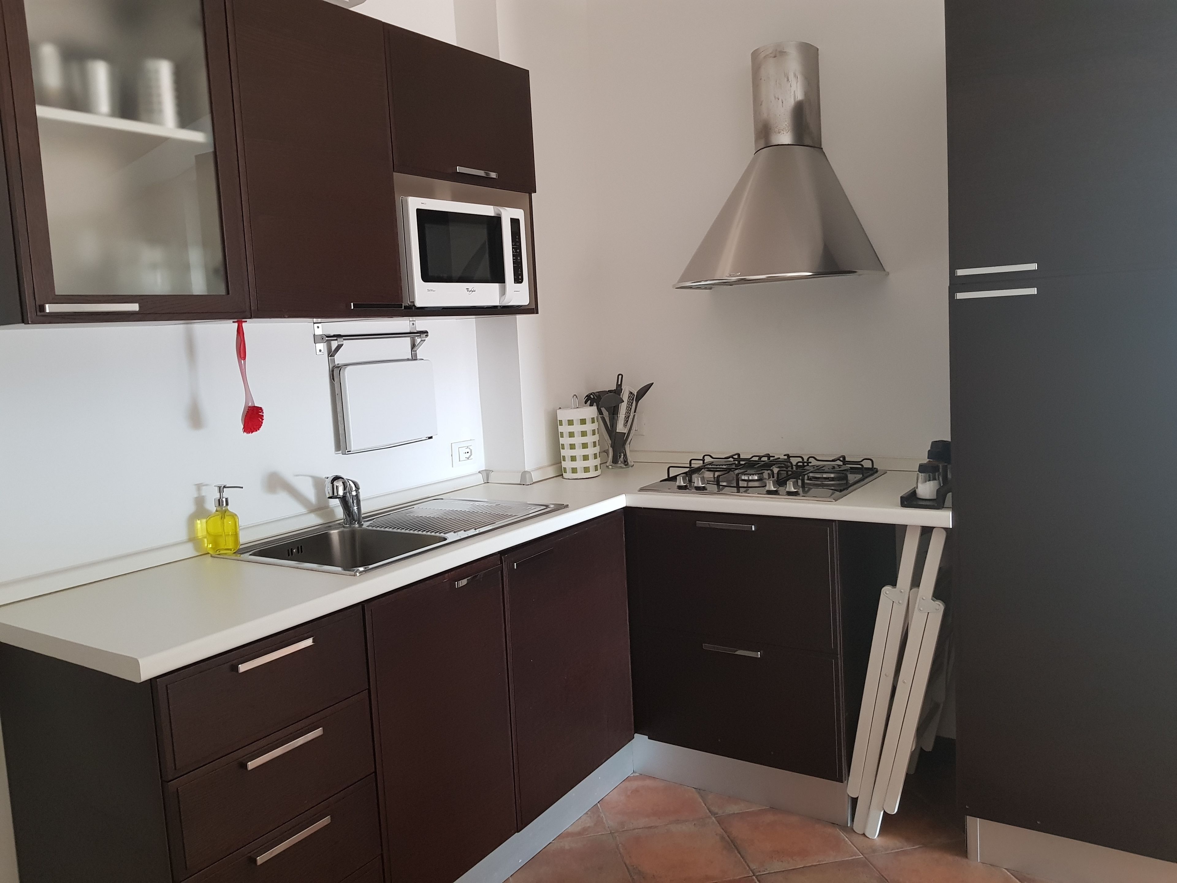 Una cocina con armarios marrón oscuro, una encimera blanca, un microondas de acero inoxidable, una estufa a gas y un cepillo de dientes rojo colgado en la pared.