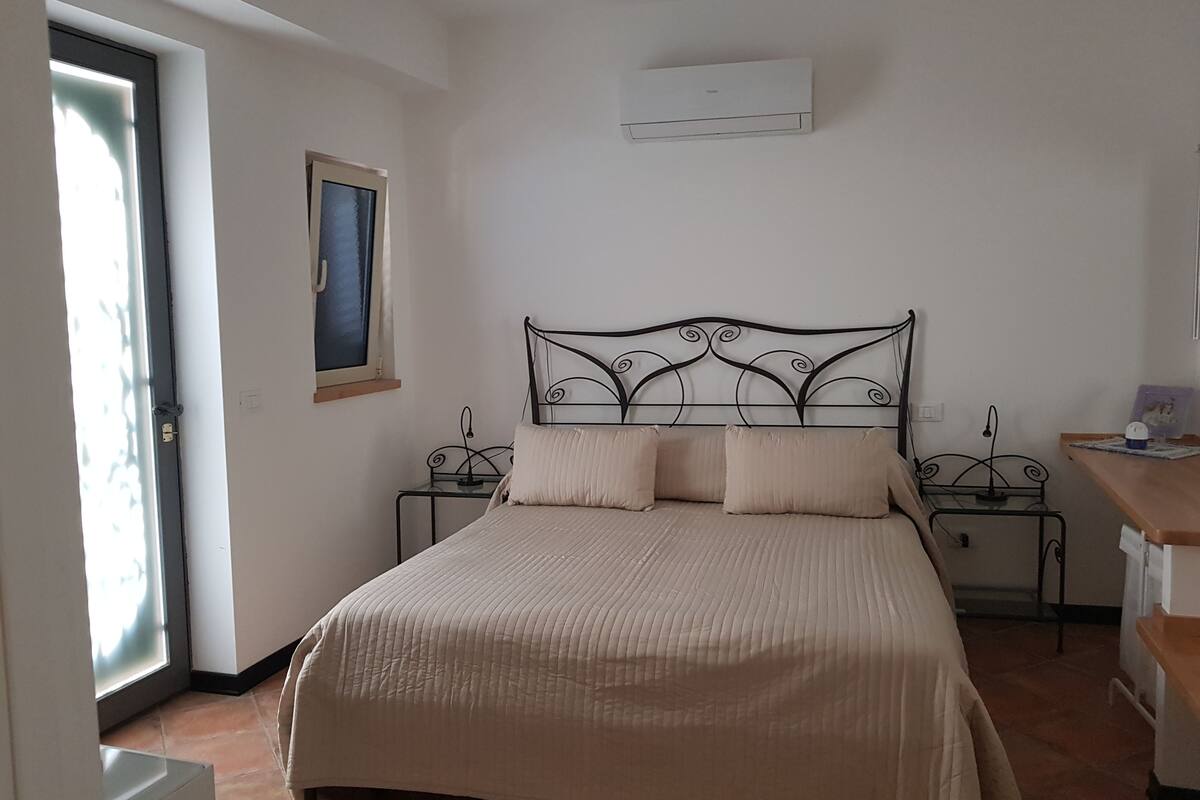 Een slaapkamer met een bed, een raam, een wandairconditioner en een nachtkastje met een lamp.