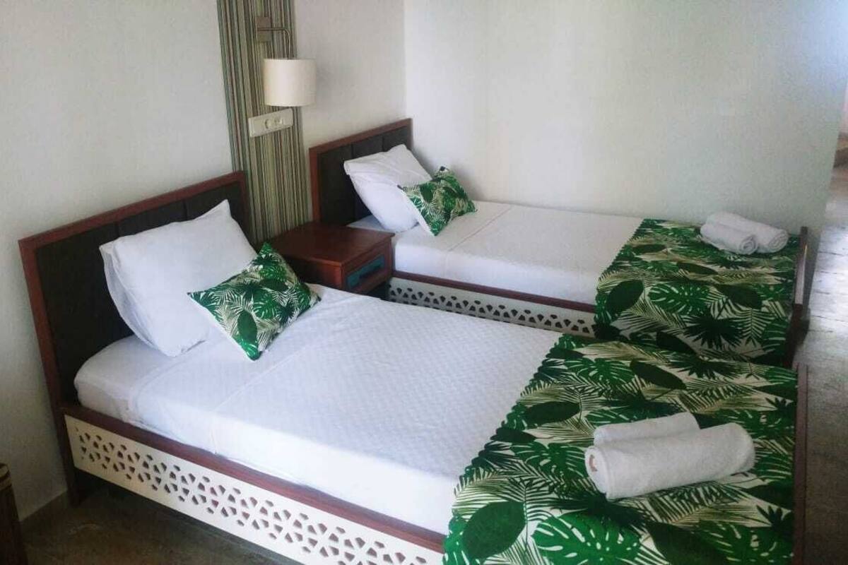 Een hotelkamer met twee enkele bedden, elk met witte lakens en kussens met een bladpatroon. Daartussen staat een klein houten nachtkastje, en erboven een wandlamp.