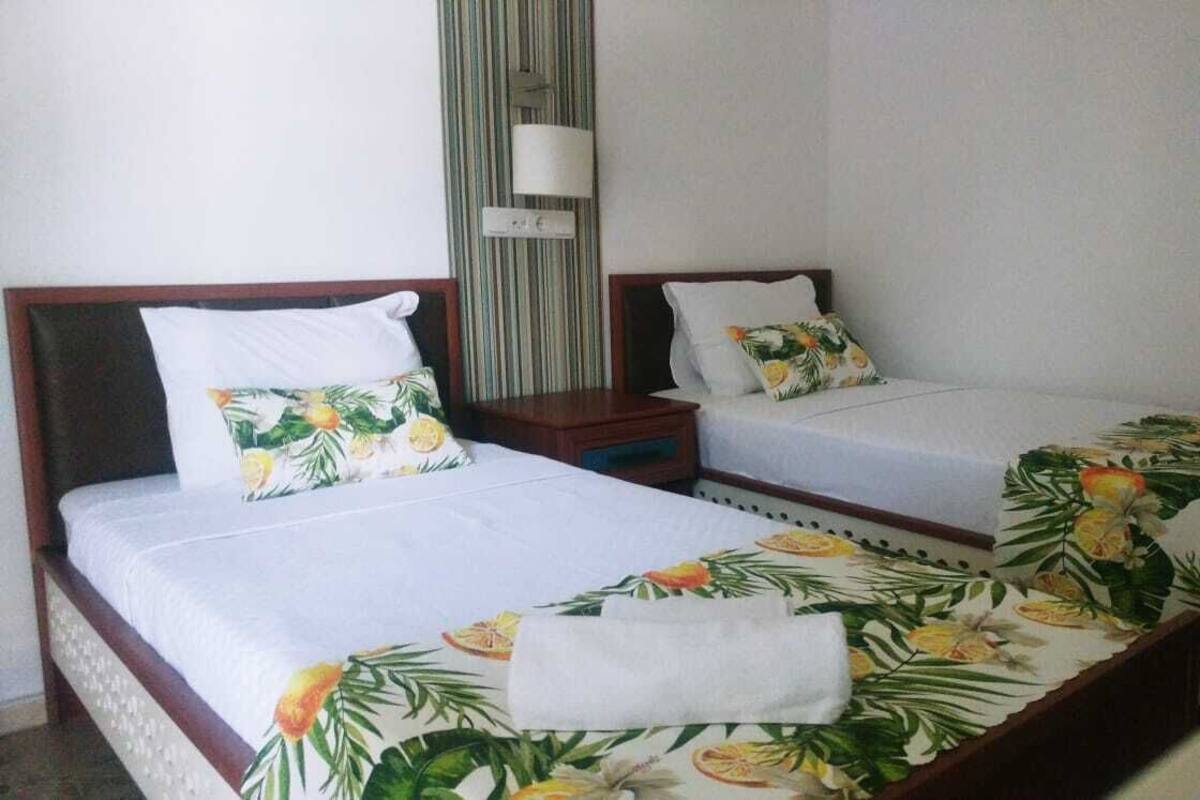 Een hotelkamer met twee eenpersoonsbedden, elk voorzien van een bloemenprint deken en een wit kussen. Tussen de bedden staat een nachtkastje met een lamp.