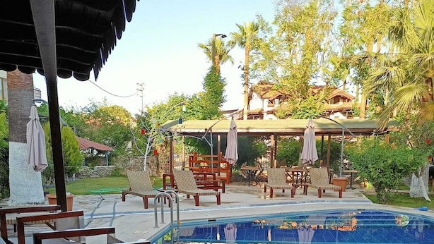 Ein Poolbereich mit Holzstühlen und -tischen, einem überdachten Sitzbereich und Blick auf den Pool, der die umliegenden Bäume reflektiert.