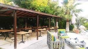 Eine Terrasse im Freien mit Holztischen und -stühlen, einem überdachten Sitzbereich und einer Palme.