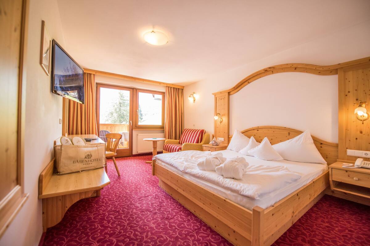 A hotel room with a wooden bed, a desk with a bag, a chair, a small table, and a TV on the wall.