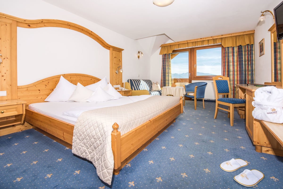 A hotel room with a large bed, a desk with a chair, a small table, and a window with curtains.