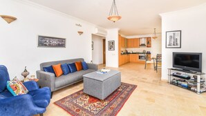 A living room with a grey sofa, blue armchair, and ottoman, a flat-screen TV, and a kitchen in the background. - Beautifully-located Large 2 bed, sea view garden apartment with shared lge pools (Kalkan)