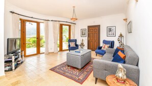 A living room with a grey sofa, a coffee table, a television, and a large window with curtains. - Beautifully-located Large 2 bed, sea view garden apartment with shared lge pools (Kalkan)