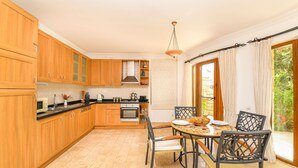 A kitchen with wooden cabinets, a stove, and a dining area with chairs and a table. - Beautifully-located Large 2 bed, sea view garden apartment with shared lge pools (Kalkan)