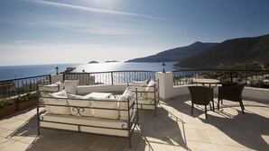 A terrace with a view of the sea and hills, featuring a bed, chairs, and a table. - Beautifully-located Large 2 bed, sea view garden apartment with shared lge pools (Kalkan)