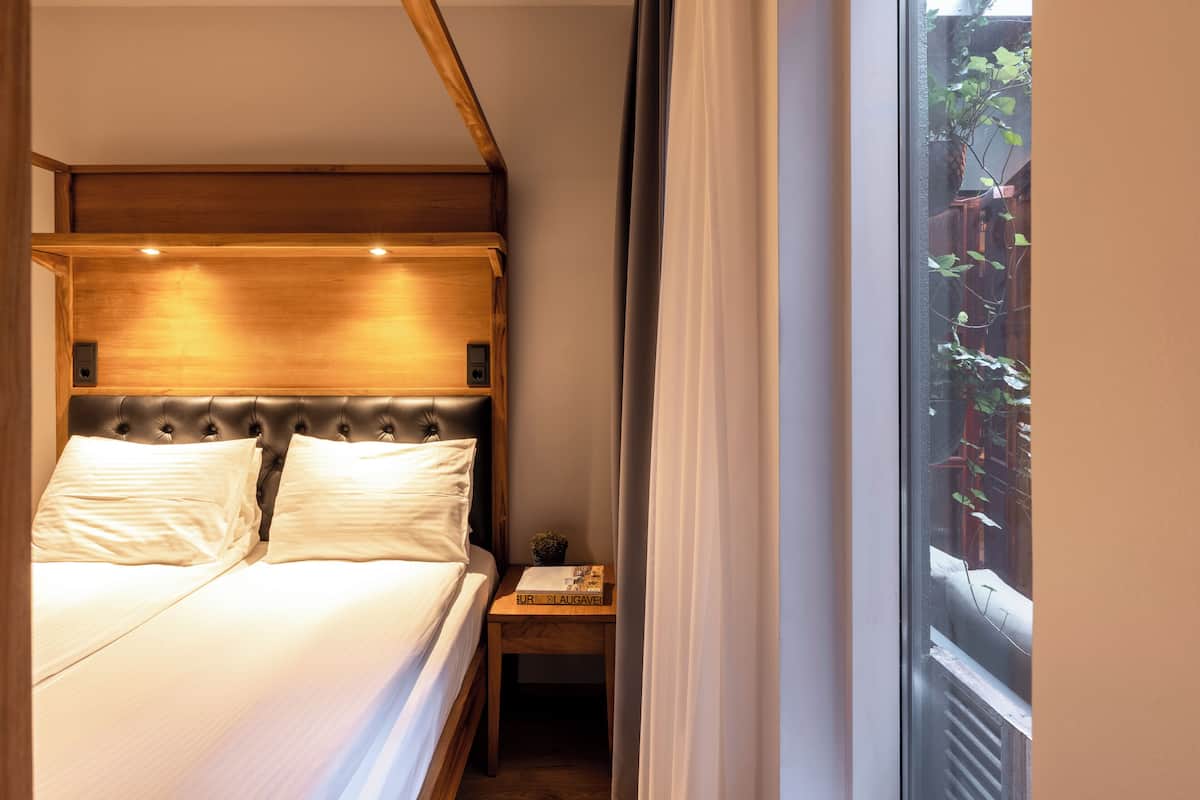 A bedroom with a wooden headboard, a small bedside table, and a window with a view of greenery outside.