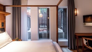 A hotel room with a large bed, a TV, and a view of an outdoor seating area through a window.