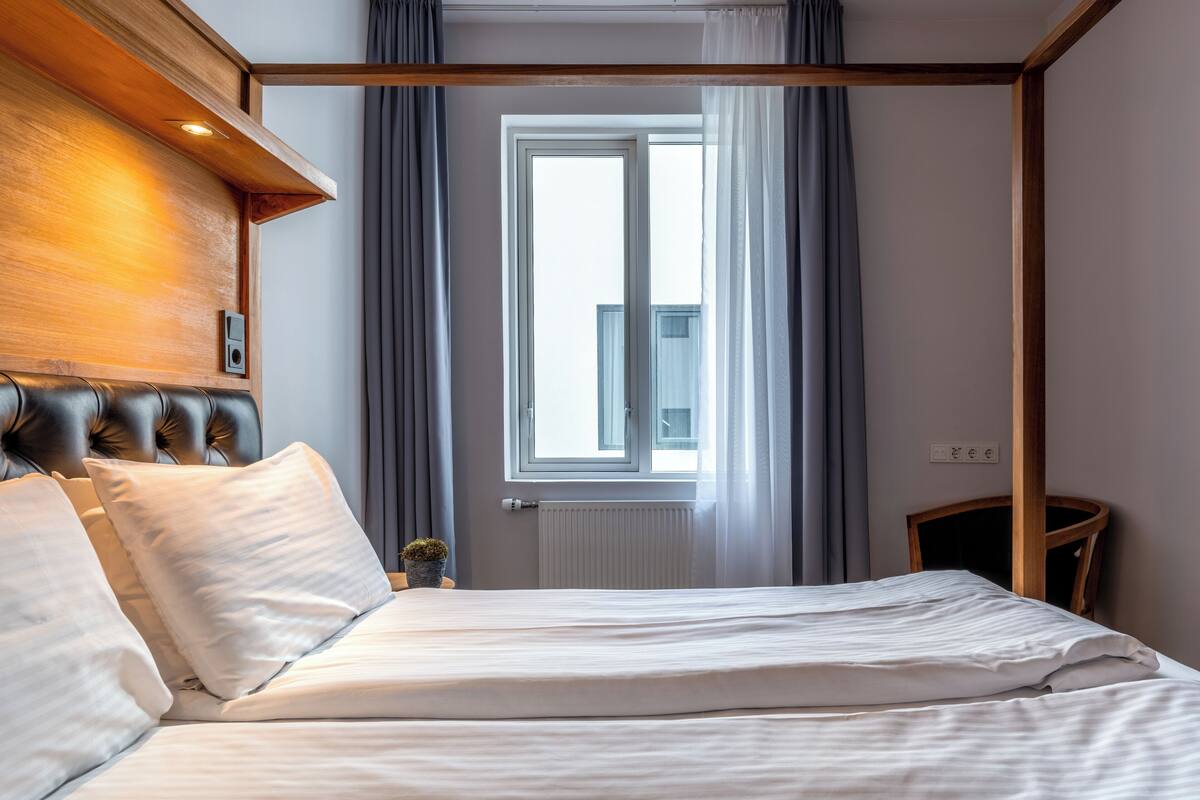 A hotel room with a large bed, a wooden headboard, a window with curtains, and a small table with a plant.
