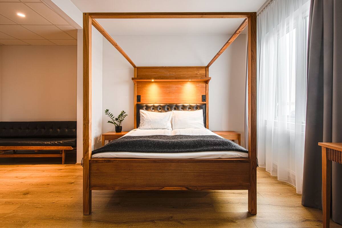 A wooden four-poster bed with a white mattress and pillows, a small bedside table with a plant, and a bench in the background.