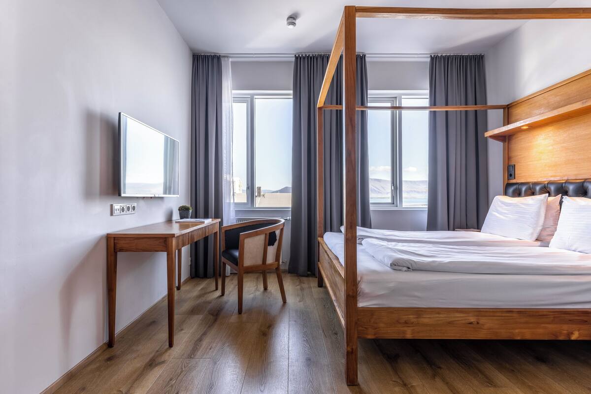 A modern hotel room with a wooden four-poster bed, a desk with a chair, a flat-screen TV, and large windows offering a view of the mountains.