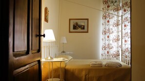 A hotel room with a bed, two bedside lamps, a small round table, a chair, a painting on the wall, and a floral curtain. - Casa Laino (Belcastro)