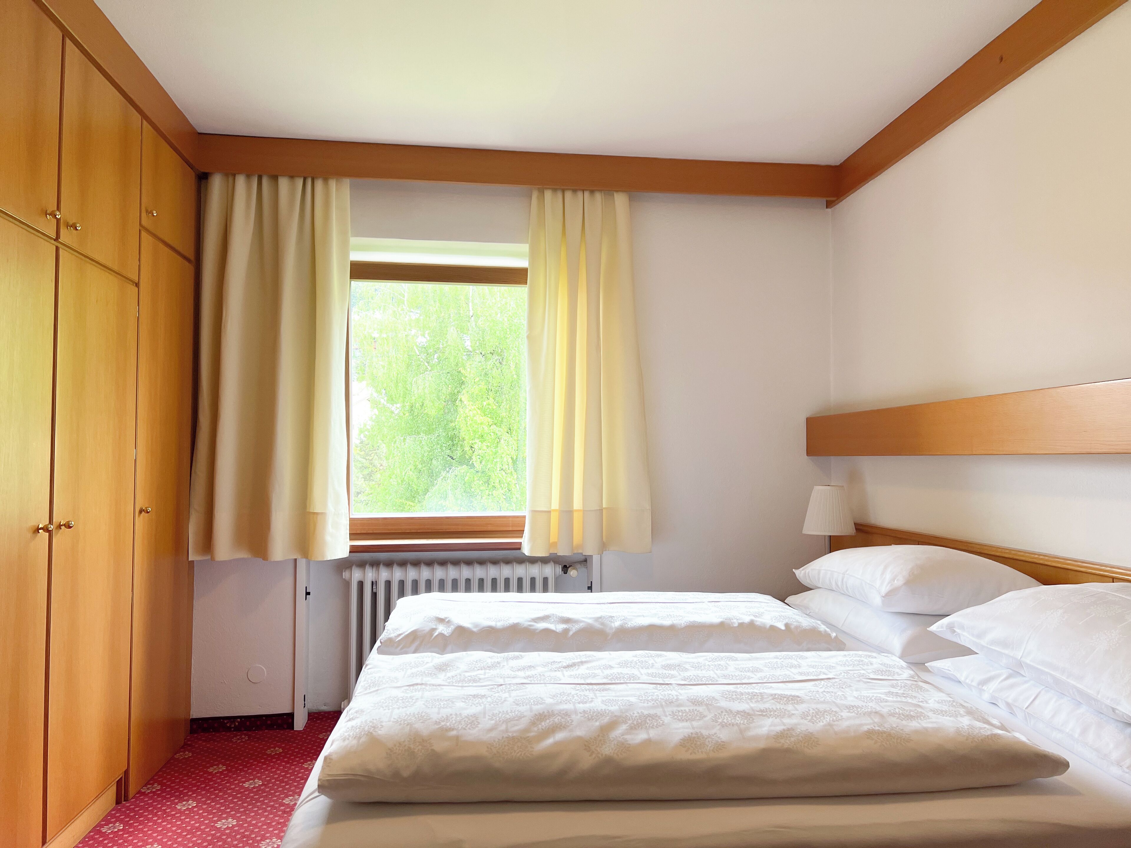 A hotel room with a wooden wardrobe, a bed with white linens, a window with curtains, and a radiator.