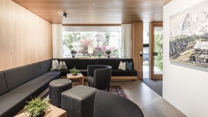 Un salon moderne avec un canapé foncé, deux poufs, une petite table et une grande fenêtre donnant sur des arbres et une montagne.
