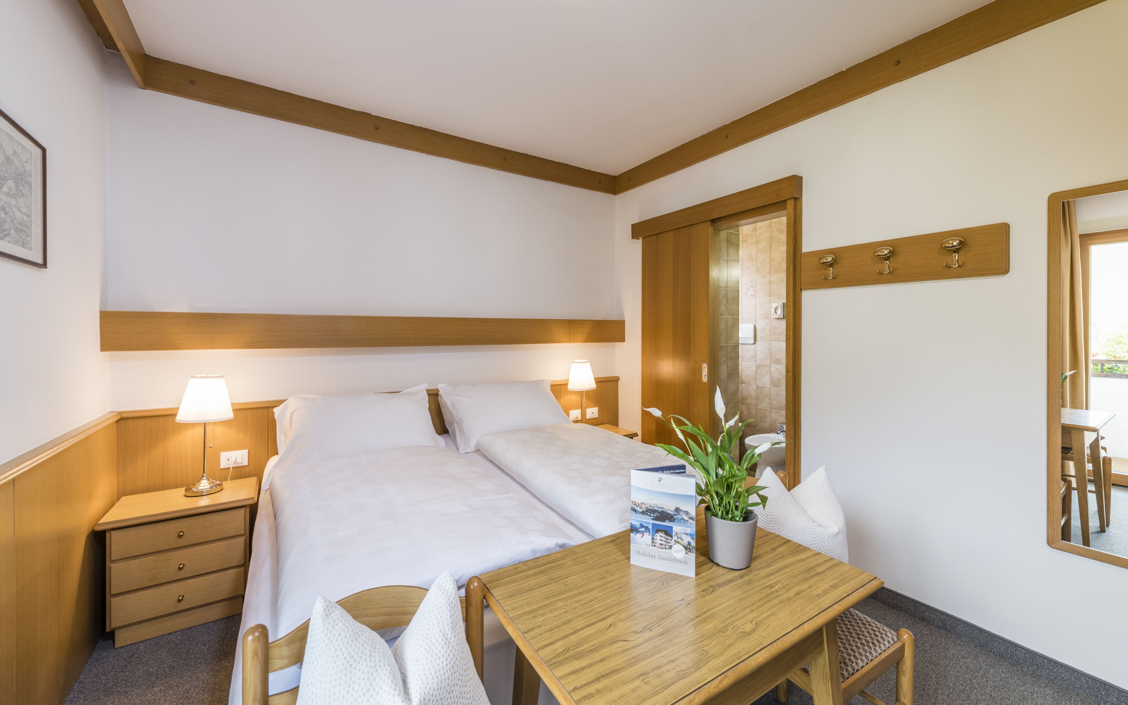 Une chambre d’hôtel avec un lit, des tables de chevet, une tête de lit en bois, une petite table avec une plante et un miroir.