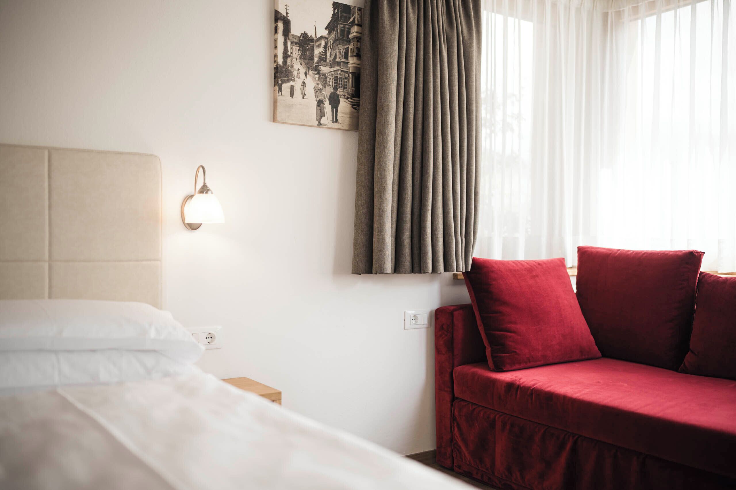 Une chambre d’hôtel avec un lit, un canapé rouge, une lampe fixée au mur et une fenêtre avec des rideaux.