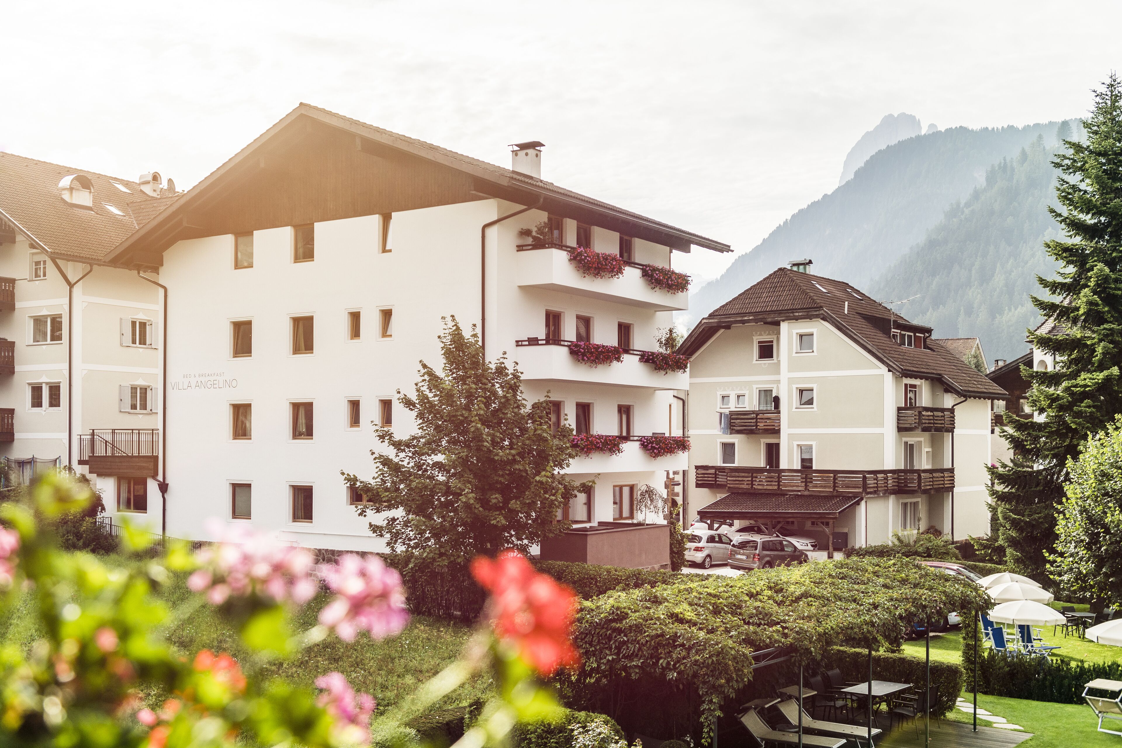 Un hotel de varios pisos con balcones, un jardín con mesas al aire libre y montañas al fondo.