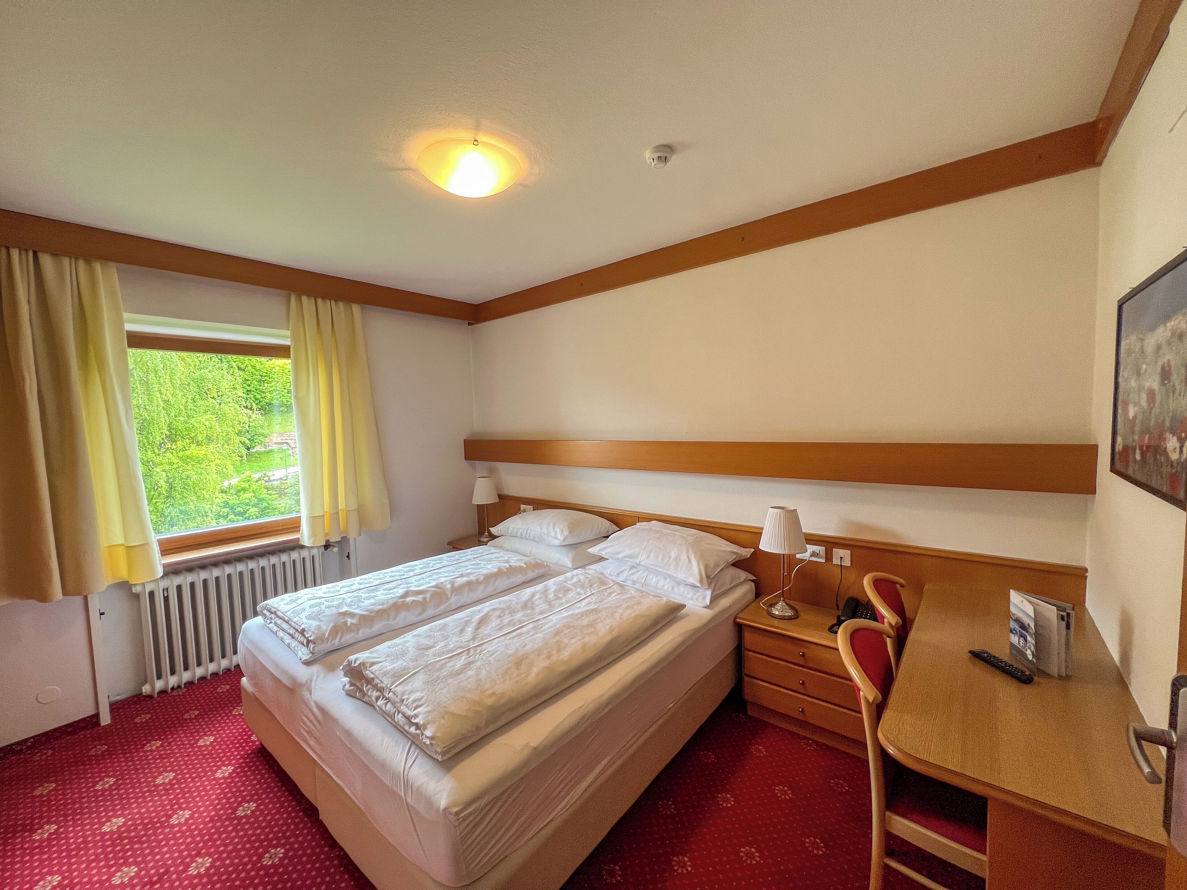 Habitación de hotel con dos camas, un escritorio, una silla, un radiador y una ventana con cortinas.