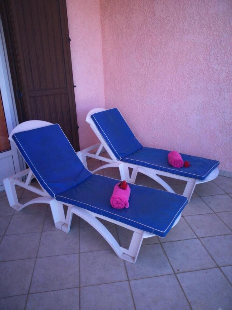 Two lounge chairs with blue cushions and pink towels, positioned on a tiled patio against a pink wall.