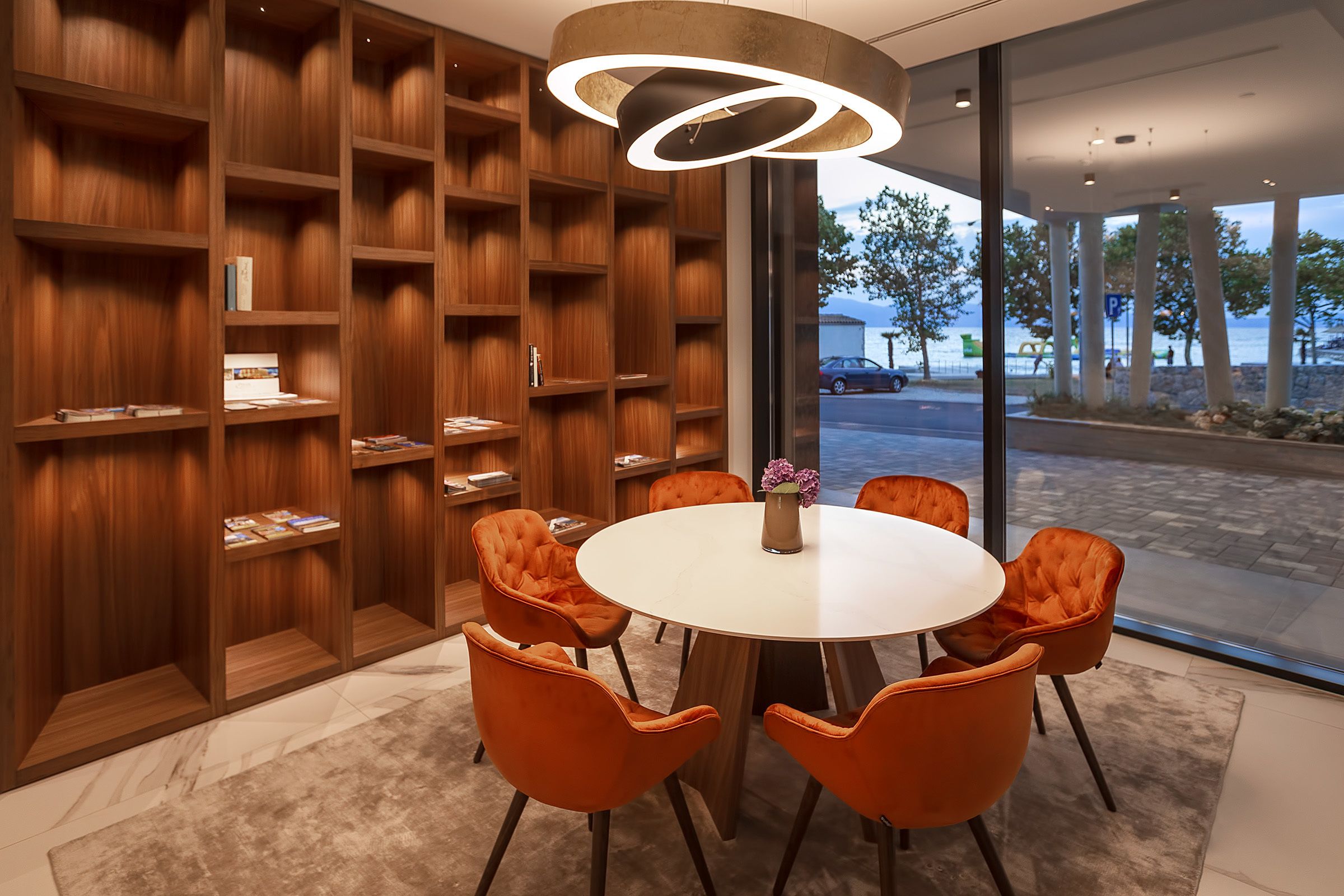 A modern interior with a round table, orange chairs, wooden bookshelves, and a view of the outdoors.