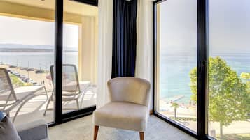 A room with a large window offering a view of the sea and beach. There is a chair and a small table in the room.