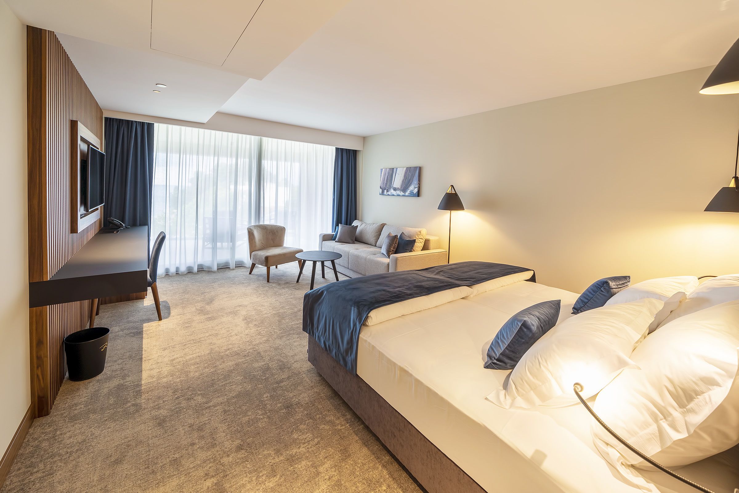 A modern hotel room with a large bed, a desk, a chair, a sofa, and a TV.