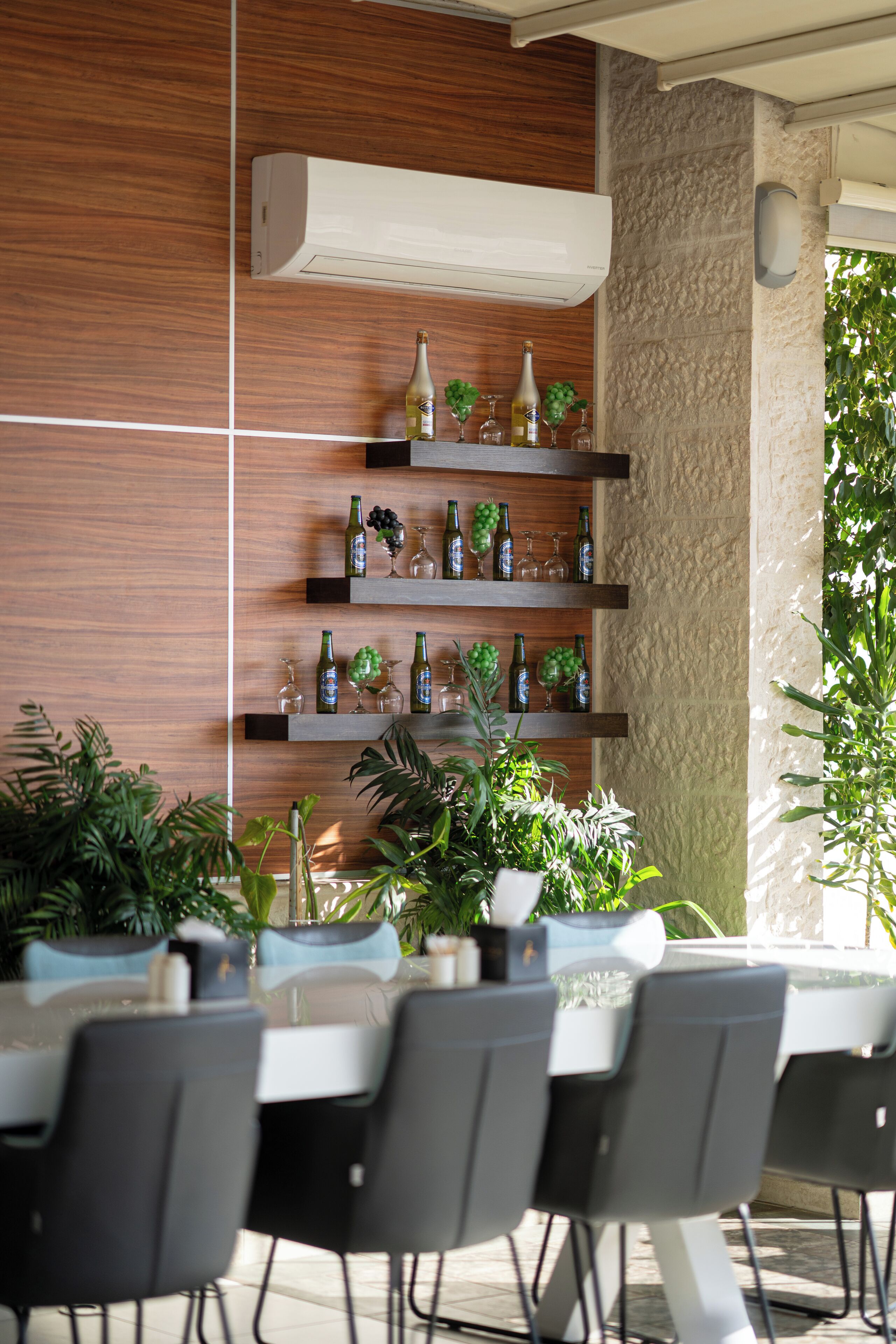 A restaurant with a modern interior, featuring a wooden wall, a stone pillar, and a shelf with bottles and glasses. There are tables and chairs arranged for dining.