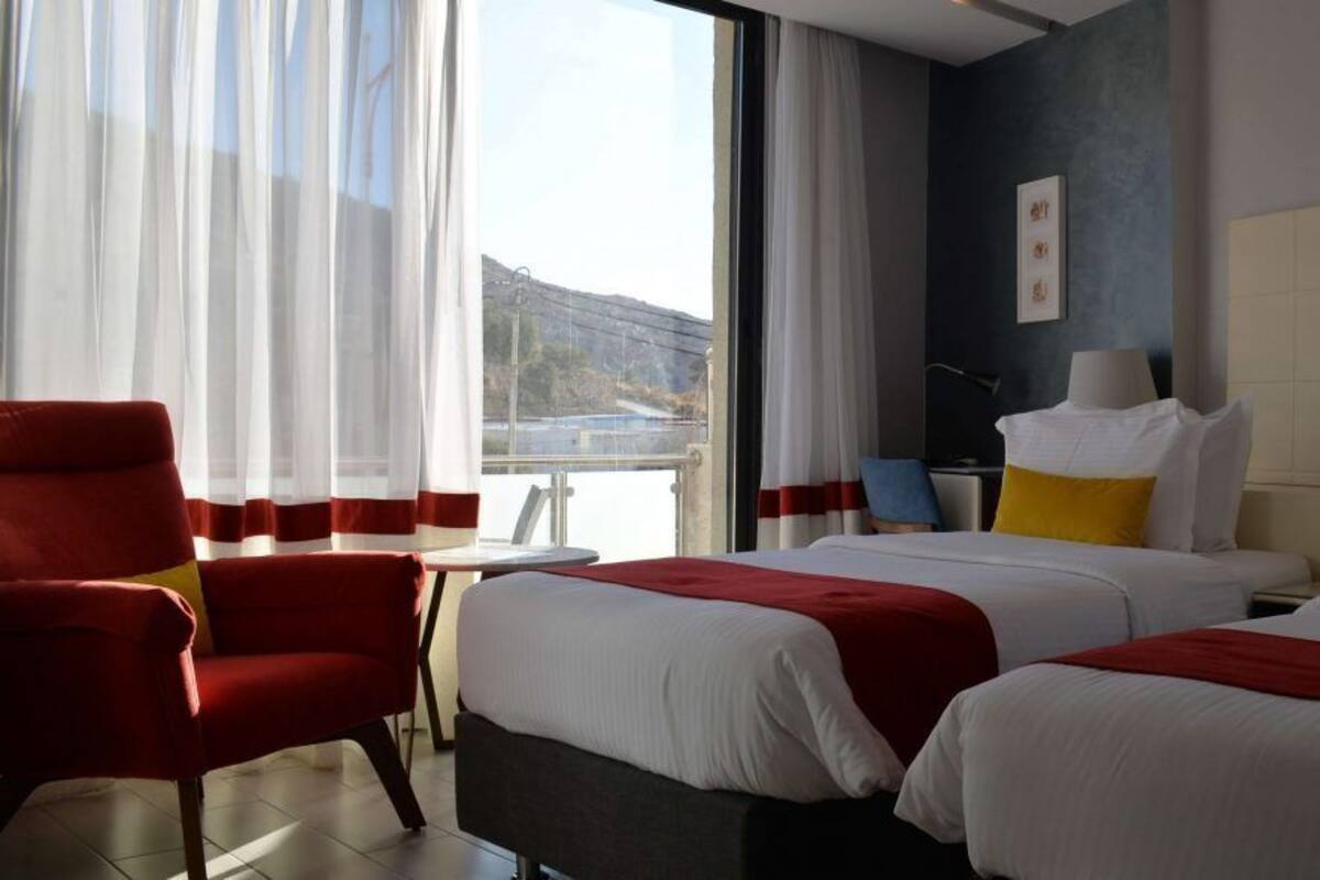 A hotel room with two beds, a red armchair, a desk, and a view of the outdoors.