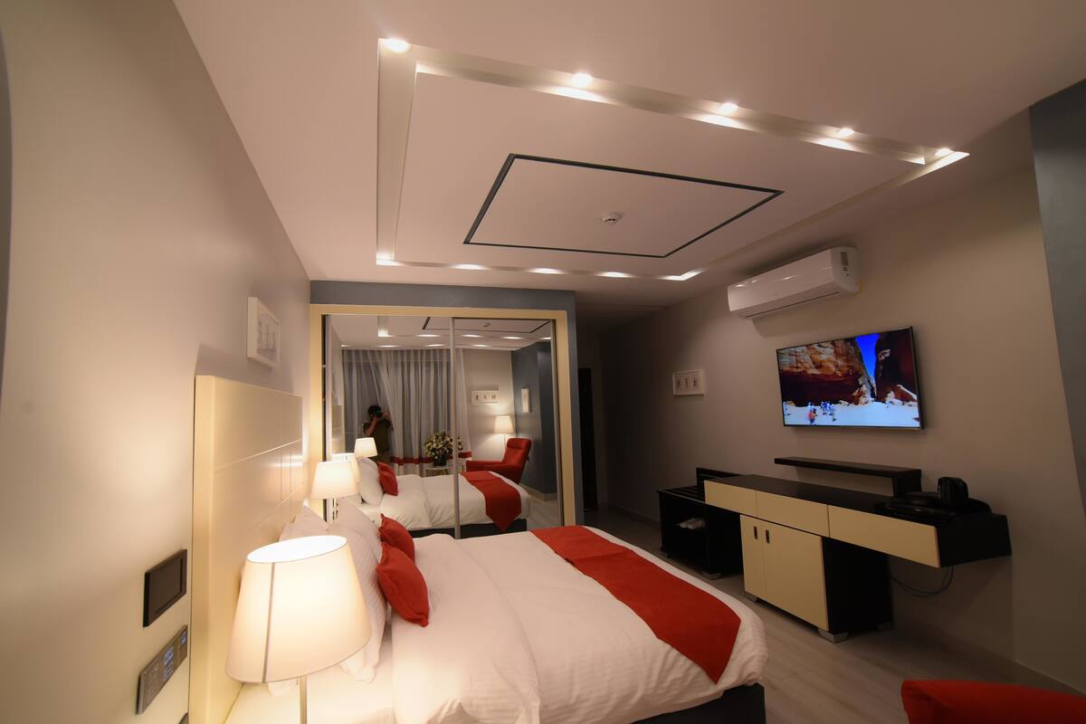 A modern hotel room with a bed, bedside table, lamp, TV, and a desk with a chair.