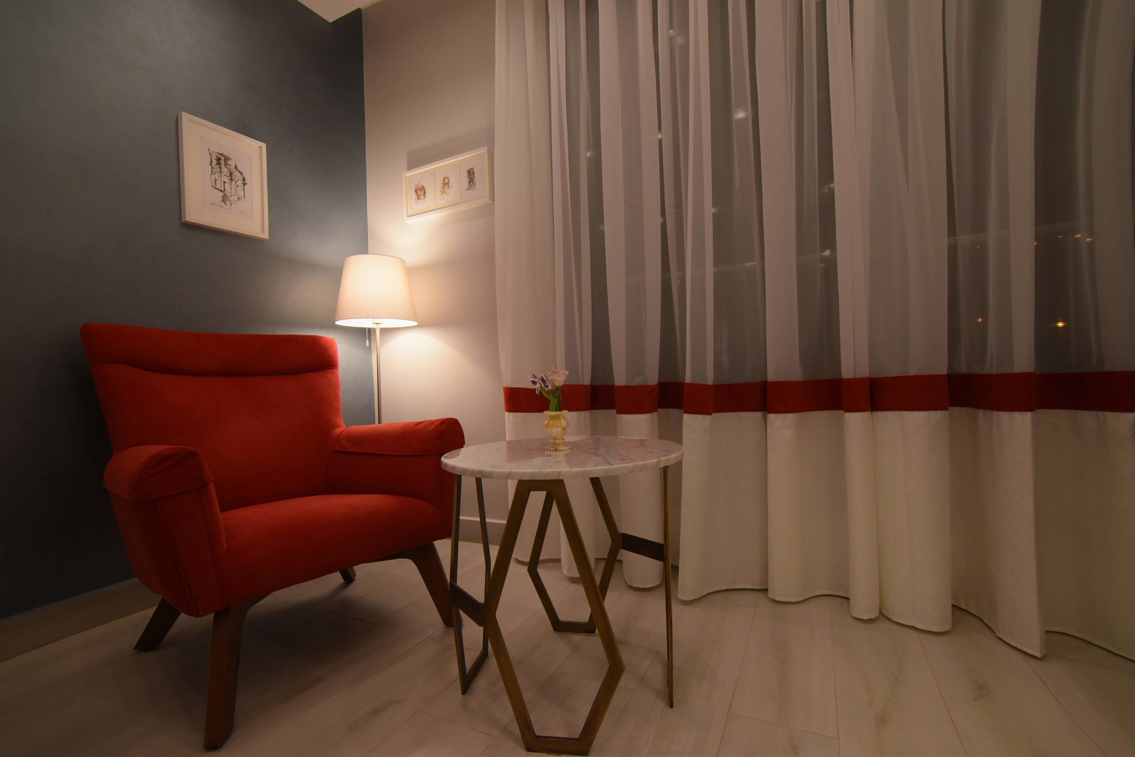 A room with a red armchair, a round table with a vase, and sheer curtains with a red stripe.