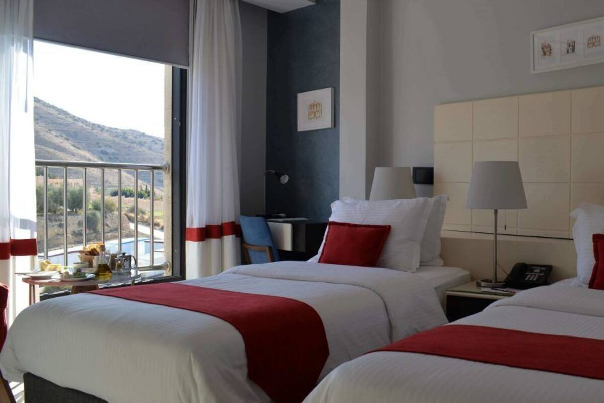 A hotel room with two beds, a desk, a chair, a lamp, and a view of the outdoors.