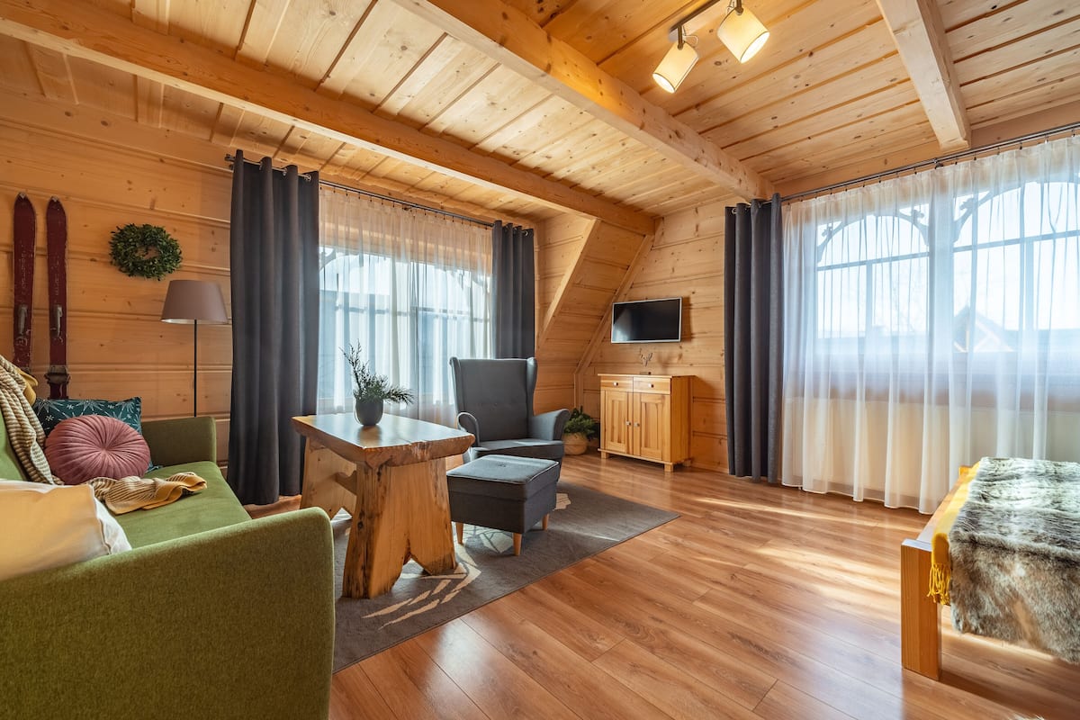 A cozy room with a wooden ceiling, a green sofa, a dark armchair, a wooden table, and a TV mounted on the wall.