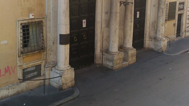 A building with three black doors, each with a sign above. There are windows on the upper floors, and a metal railing on the balcony. - Domus Del Corso Spagna (Rome)
