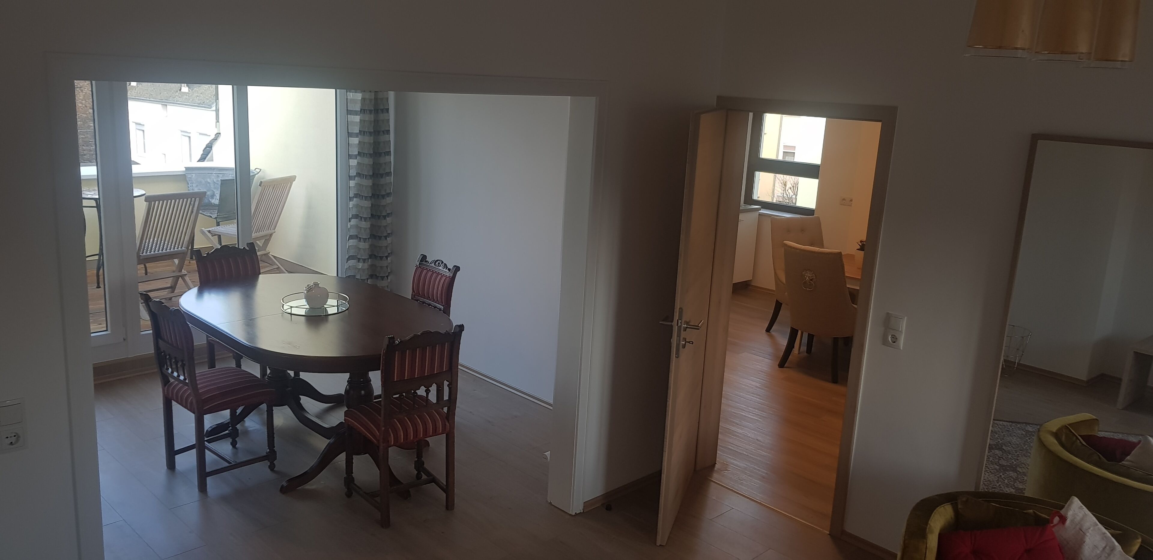 A dining area with a table and chairs, a view through an open door to a kitchen, and a window with curtains.
