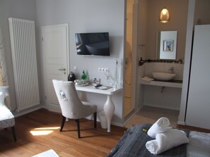 A hotel room with a wooden floor, a white desk with a chair, a television mounted above the desk, a bathroom with a sink and mirror, and towels on the bed.
