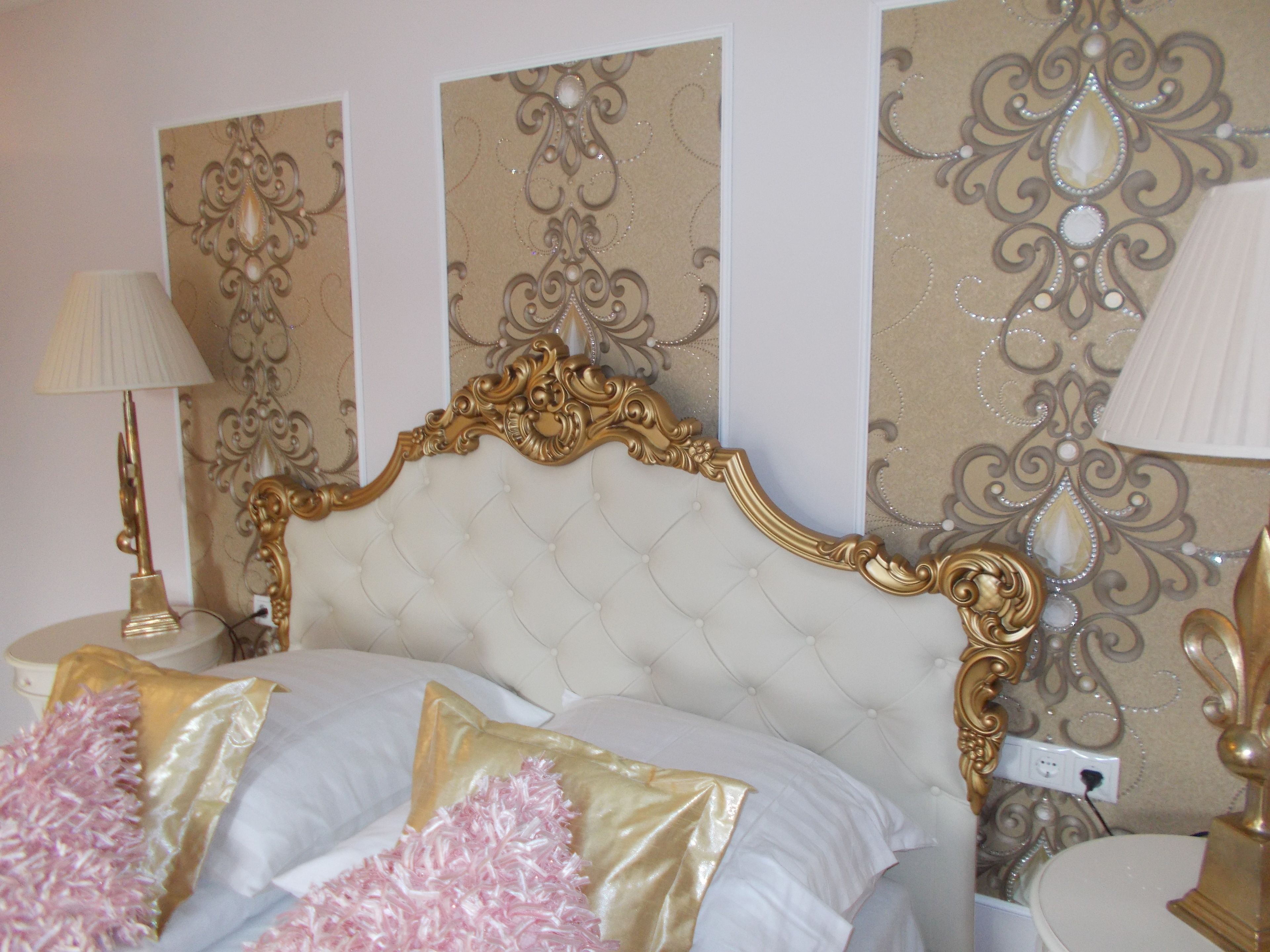 A bedroom with a tufted headboard, decorative wall panels, a bedside table with a lamp, and a bed with white and gold bedding.
